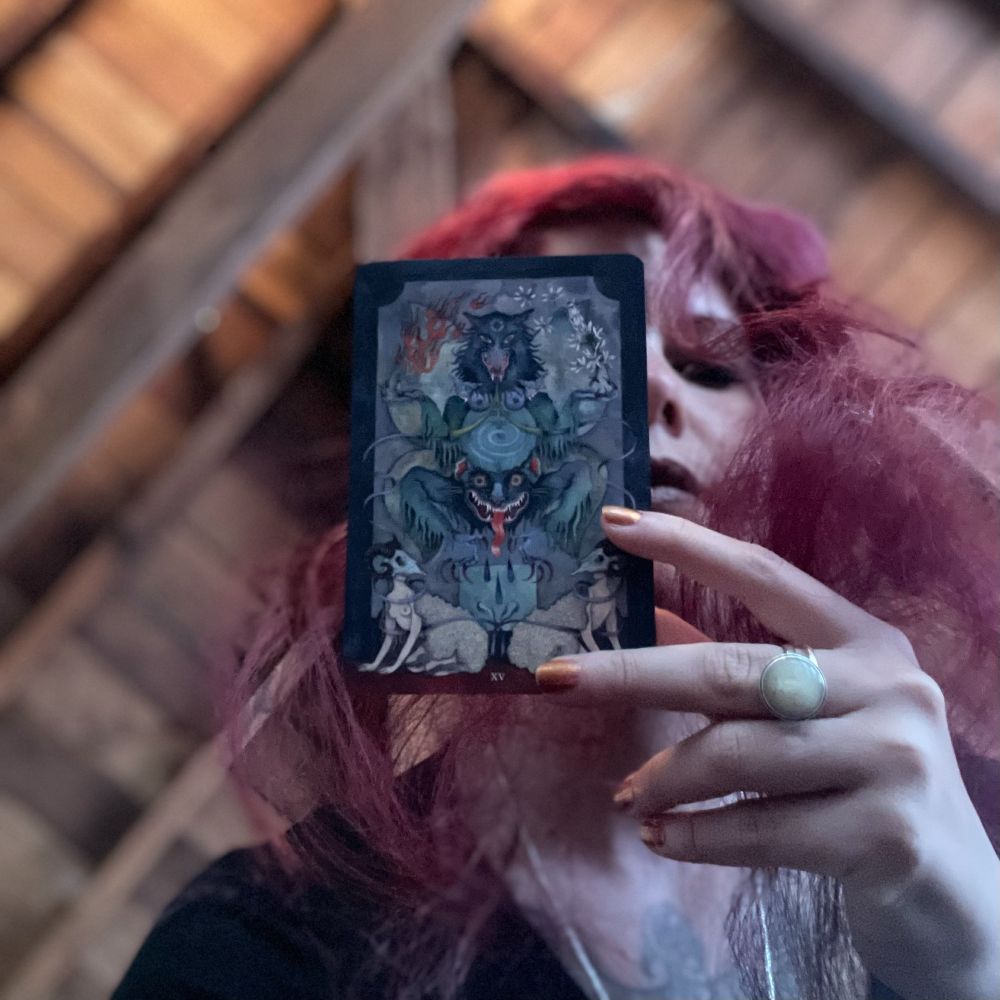 A person with long, wavy red hair holds up a tarot card in front of their face, partially obscuring their features. The card shows a vivid, surreal depiction of the Devil with beastly faces, claws, and flames. The background is an angled wooden ceiling, and the lighting is soft and moody. The person wears a large round ring and has copper-colored nails.