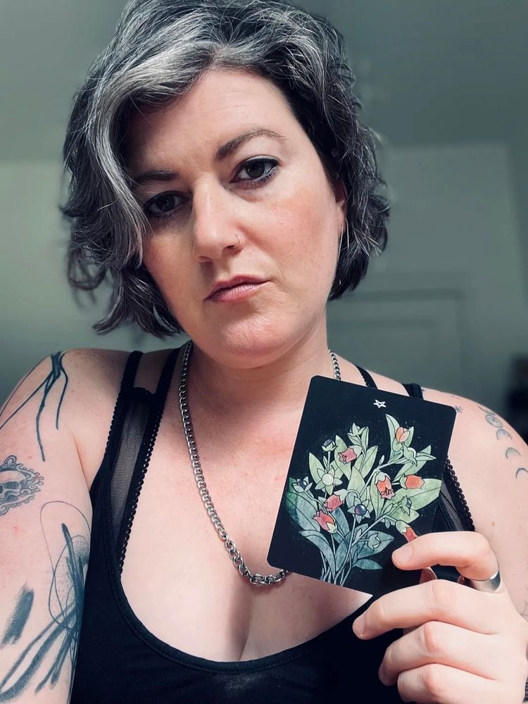 Meg, who has short wavy gray and brown hair, wearing a black tank top and a silver chain necklace, holds up the Devil card from Botanical tarot featuring an illustration of flowering plants with green leaves and pink blossoms. Meg has tattoos on both arms and a nose ring, and gazes directly at the camera with a neutral expression. The background is softly lit and out of focus.

