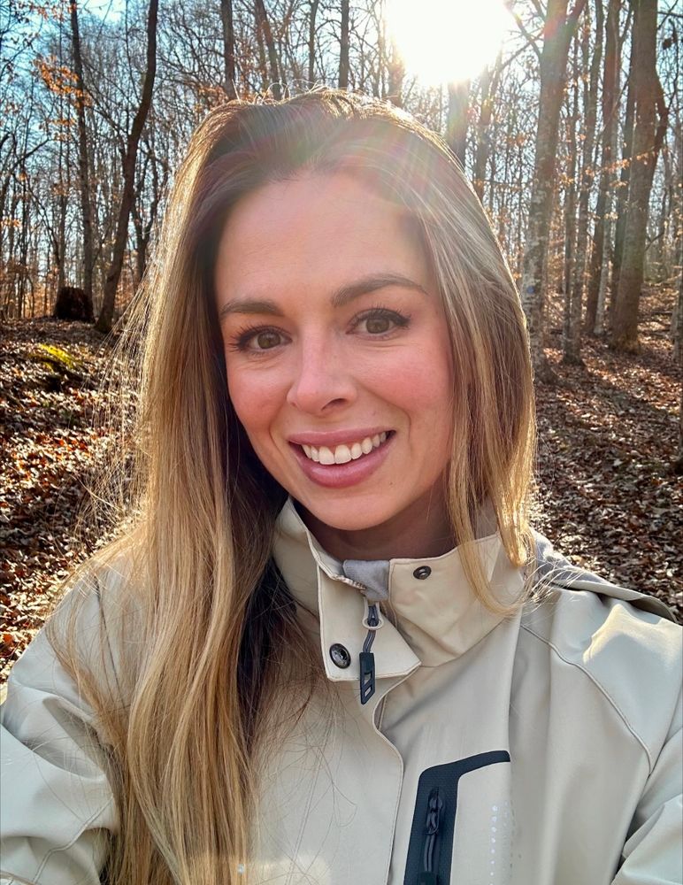 A woman with long, light brown hair smiles at the camera in a forest. She is wearing a beige jacket with a black zipper detail. The sun shines brightly behind her, casting a warm glow and creating a lens flare. Bare trees surround her, and fallen leaves cover the forest floor, suggesting a late autumn or early winter setting.