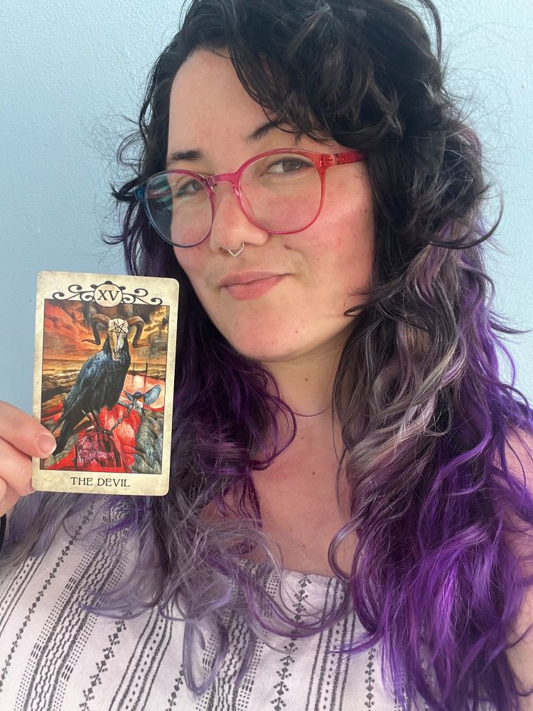 A person with long curly hair dyed black, purple, and silver poses against a light blue wall. They wear red-framed glasses, a nose ring, and a white and black patterned top. They hold up a tarot card—The Devil—from the Crow Tarot deck, which features a black crow, chains, and a fiery background.