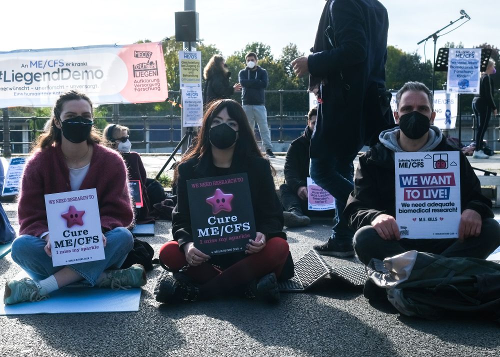 Three people are sitting cross-legged on the ground at a protest, holding signs. The person on the left has a sign reading, ‘We need research to cure ME/CFS. I miss my sparkle.’ The person in the middle holds a similar sign, and the person on the right has a sign saying, ‘We want to live! We need adequate biomedical research! M.E. kills.’ All are wearing masks. A protest banner is visible in the background, along with other protesters and signs, creating a peaceful demonstration scene.