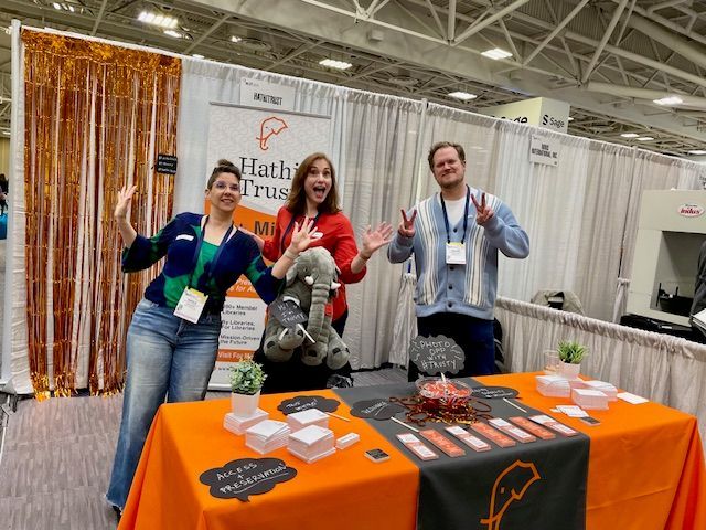Three people standing in front of a table with an orange tablecloth at a trade show. The middle person is holding a large stuffed elephant.