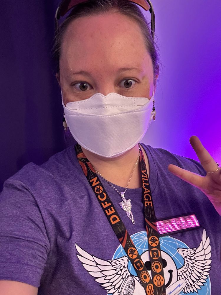 Selfie of the author who is wearing a KF94 mask, PCB earrings, Evenstar necklace, DEFCON village lanyard, and a blinky name badge that is scrolling the name “GattaKat”