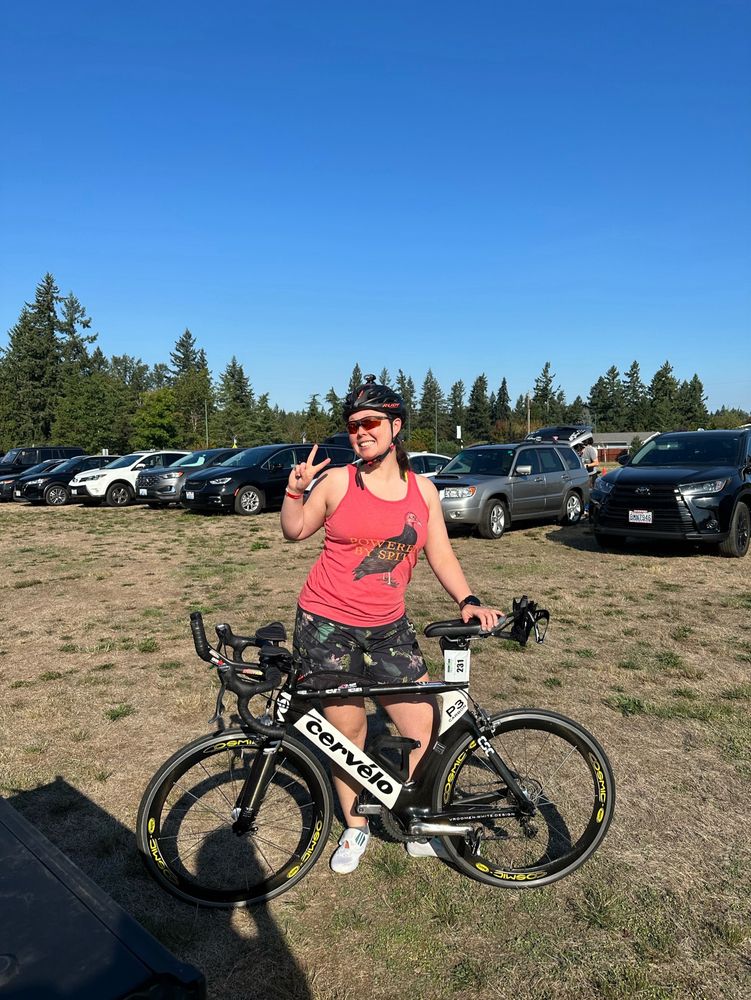 A woman stands behind a time trial triathlon bike wearing shorts and a red tank top. The tank top is from Effin Birds and says “Powered By Spite” on top of a turkey.