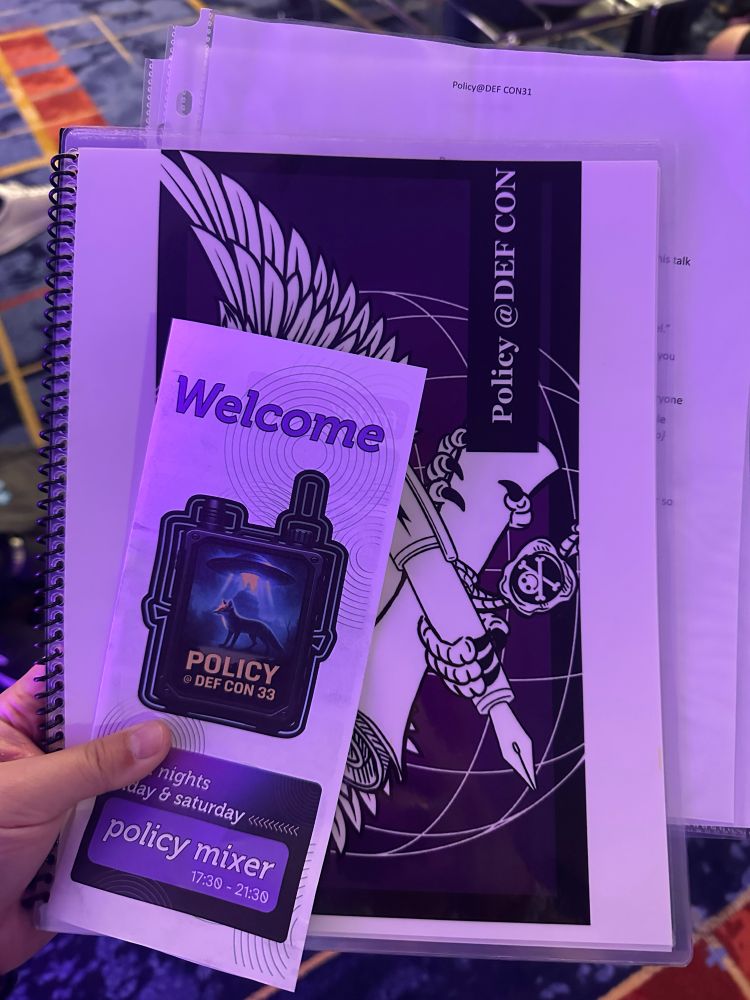 A trifold flyer that says welcome as well as “policy DEFCON 33” on top of a spiral bound notebook that has some pieces of paper sticking out that say “ROOM HOST SCRIPT”