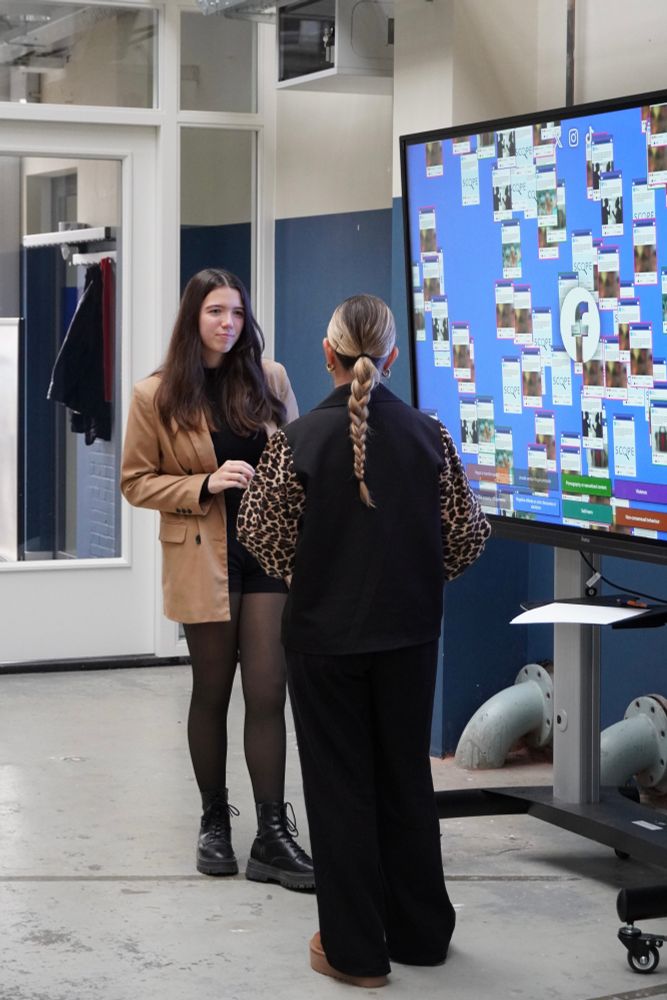 Twee mensen die praten met elkaar, met rechts een groot scherm die gemodereerde posts op Facebook toont.