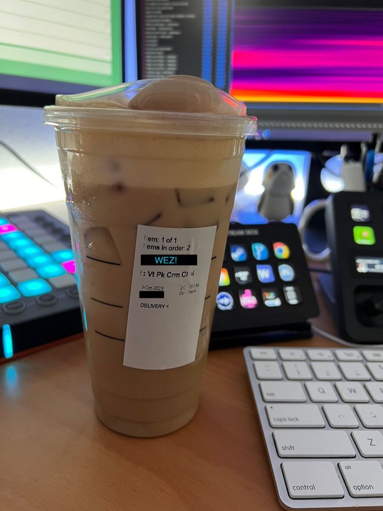 Closeup photograph of a venti pumpkin cream iced chai latte from Starbucks, with an order name edited to obscure the name of the ordered, and replaced with the text “WEZ!” The plastic cup is sitting atop a desk with an apple keyboard, two Elgato Stream Decks, a Novation Launchpad Pro, and a Star Wars Porg Funko Pop Vinyl displayed nearby.