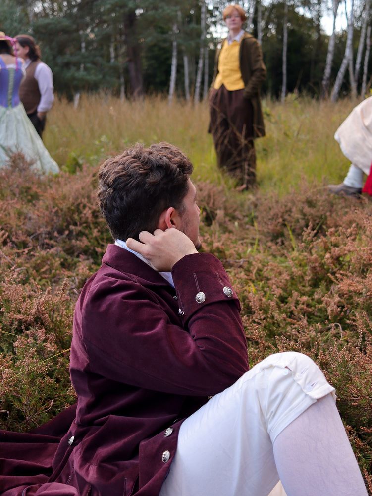 Photo of a man lounging around outdoors dressed like a gentleman from the late 18th century - purple frock coat, white floral waistcoat, white breeches. 
