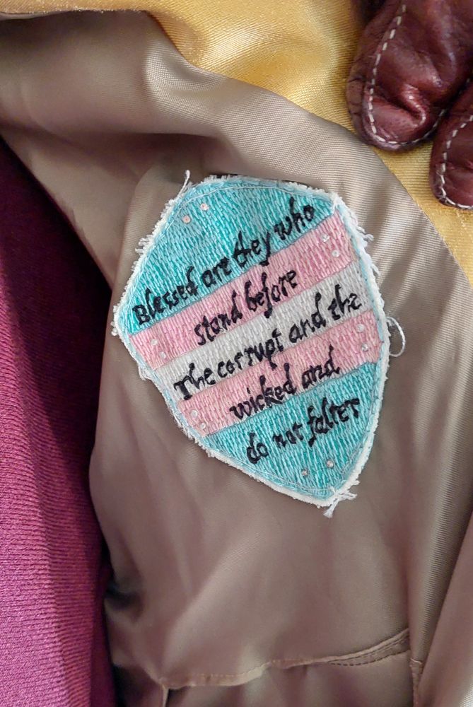 Embroidered trans flag shield with text "blessed are they who stand before the corrupt and the wicked and do not falter"