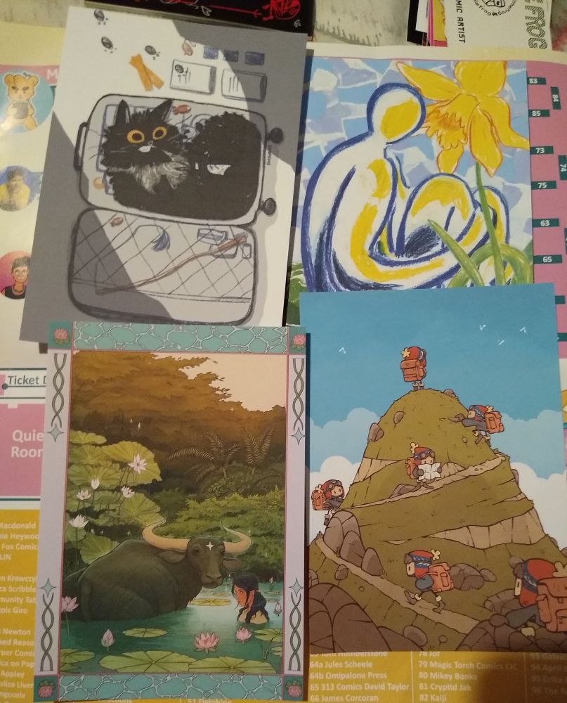 souvenier postcard set for the event, featuring art by @bhankadraws.bsky.social 
@bellerowan.bsky.social
 @francoisgiro.bsky.social
 and @mackjagee.bsky.social
top left, image of cat in a suitcase
top right, image of person sitting beside massive yellow flower.
bottom left, image of water buffalo, with a woman beside it in the water
bottom right, image of character repeating to show motion as it sumits a hill
 