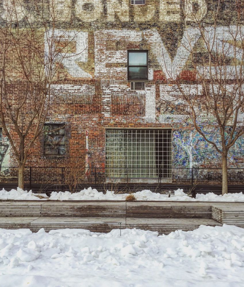 A stadium-style wooden bench partially buried in snow. Behind the bench stand bare saplings and a brick wall with windows and vintage advertising painted on it 