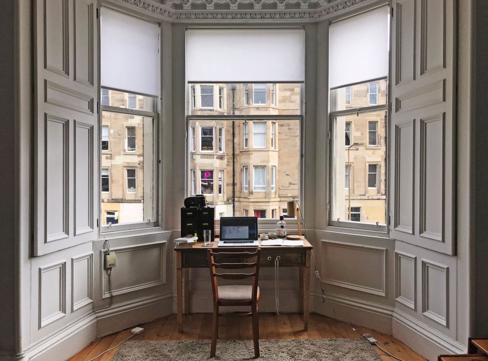 A large, bright bay window overlooking the street in a limestone Victorian row house. A wooden desk stands before the window so that, when seated, one can take in the view