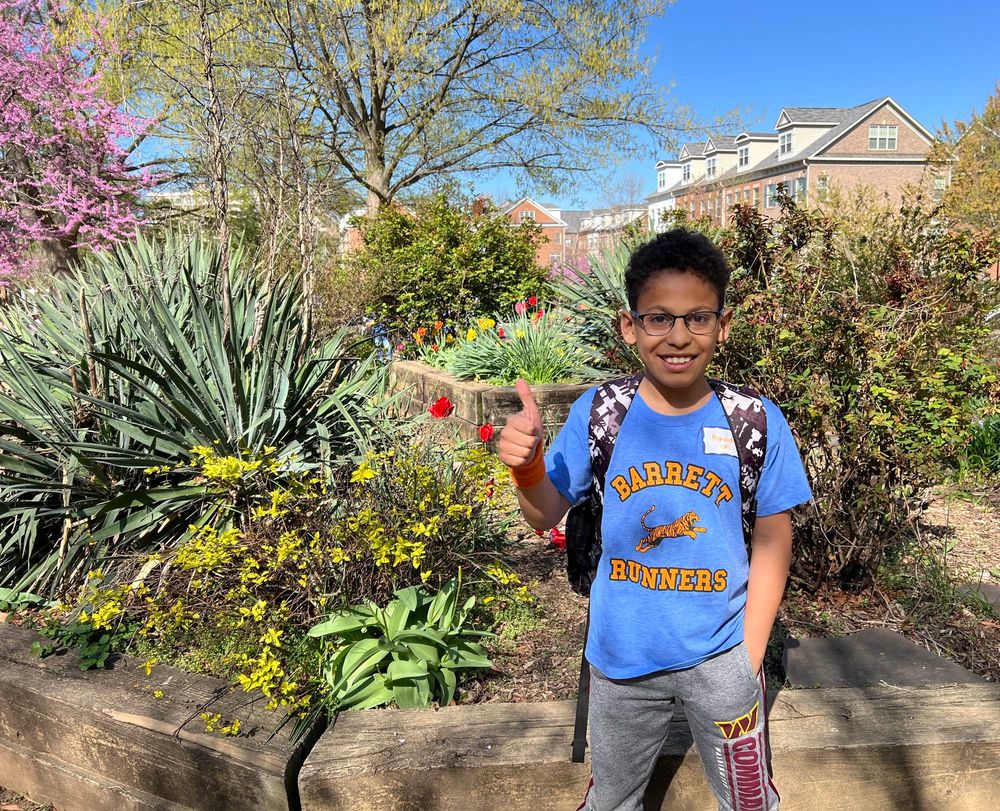 student wearing a Barrett runner blue T-shirt stands in front of the peace garden with colorful flowers.