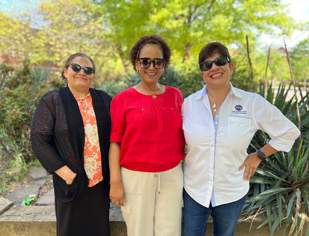The office staff is smiling and wearing sunglasses outside in front of the school Peace Garden