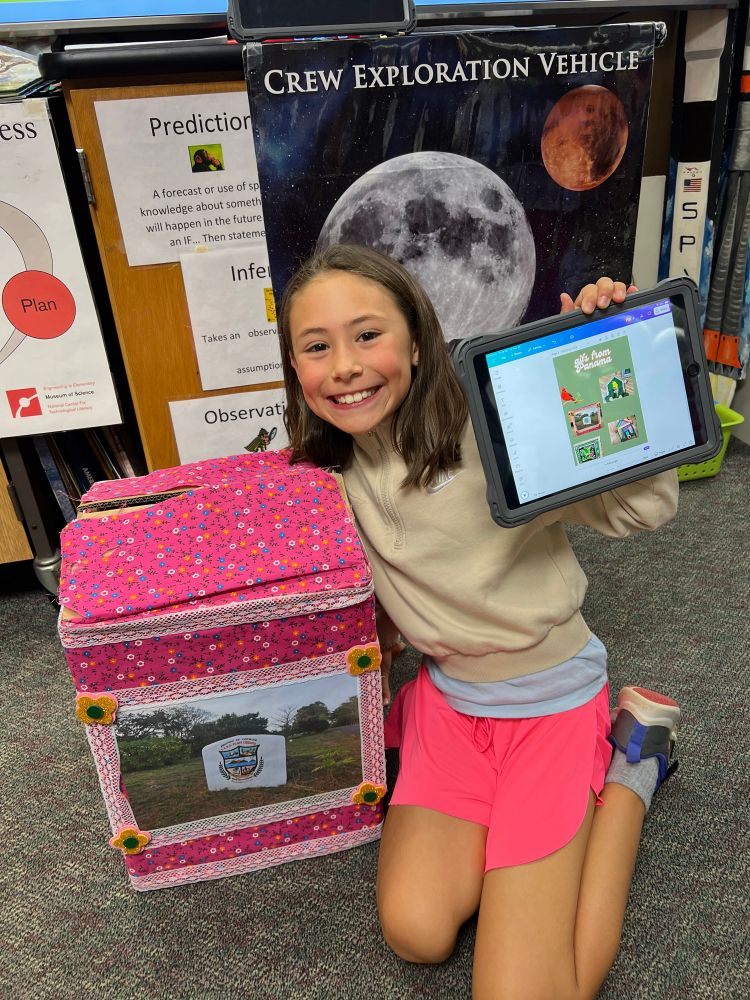 Student by a box wrapped in pink fabric holding an iPad show a creation she made in Canva with the bird artwork sent from Panama 