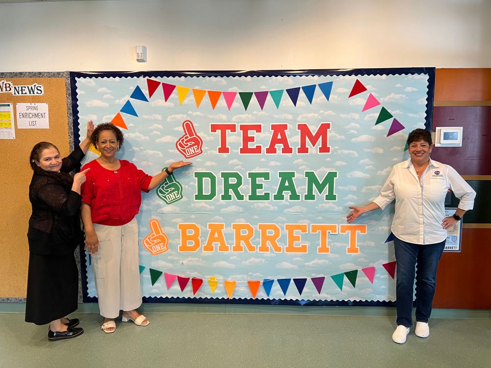 Office staff inside the school lobby by banner “1 Team 1 Dream 1 Barrett”