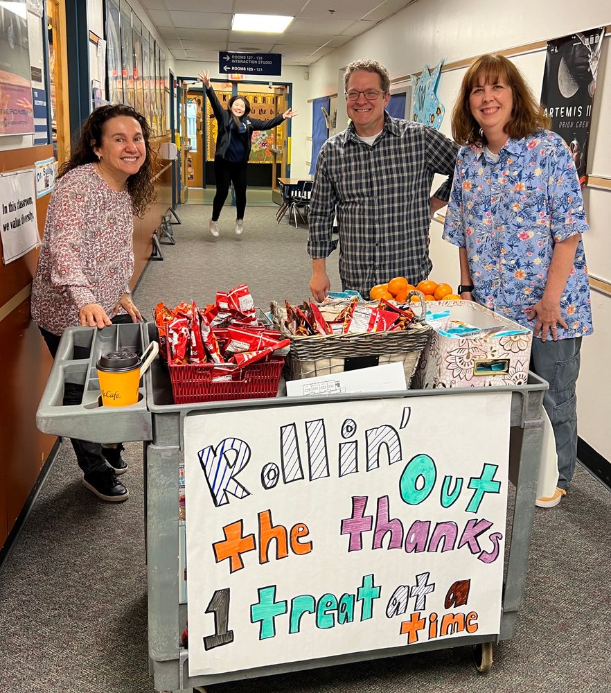 Teachers and Assistant Principal by a cart with a sign saying “Rollin out the thanks 1 treat at a time”. The principal is jumping in the air in the background 