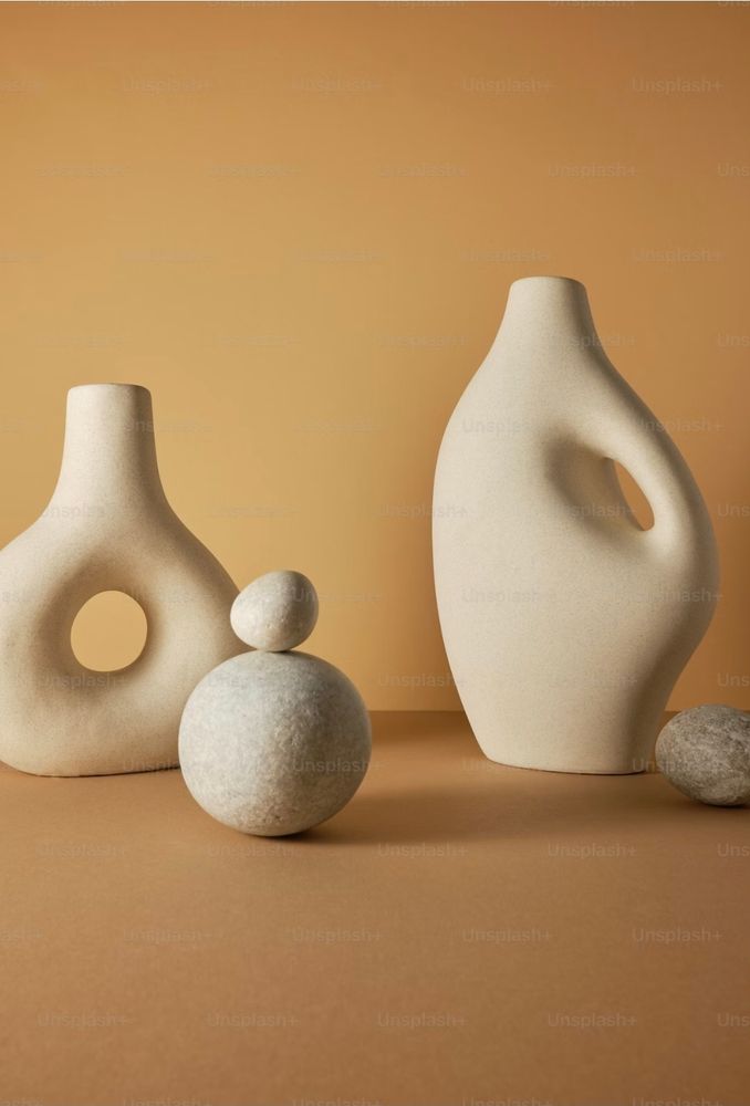 A still life photograph of ceramic jars and some rocks. On the left is a round jar with a decorative hole through the middle to the right and a little in front is 2 rocks one large round rock and a smaller one balanced on top. Then there is a larger jar that is curved and the handle is built into the curves. And beside it to the rifht is another small rock.
