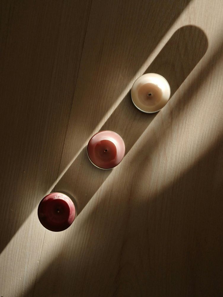 A picture of 3 candles one dark red, one pink, and one white in a line, casting shadows onto the one behind it, sitting in a direct beam of light on a wooden table.