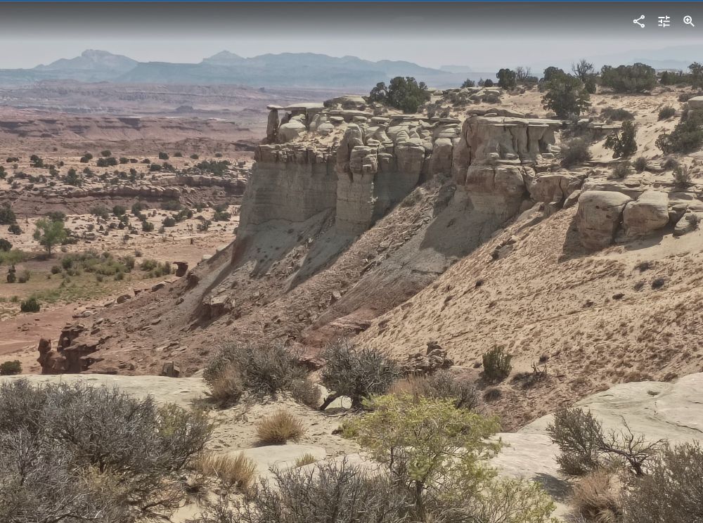 Photo off the highway somewhere in southwestern Utah, looking at a rocky mesa with a few scrub oaks dotting the sand. In the far background are some grey and distant mountains