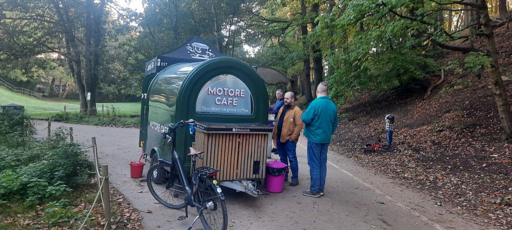 A covered trailer in a park, with the name "Motore Cafe" and the tagline "You deserve good coffee". There is a bicycle parked beside it, and a queue of people waiting for their drinks. There are lots of trees next to the path.