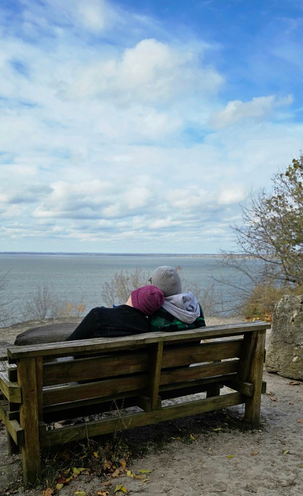 A picture of Alec and me on a chilly day, sitting on a bench on a cliff overlooking a lake.