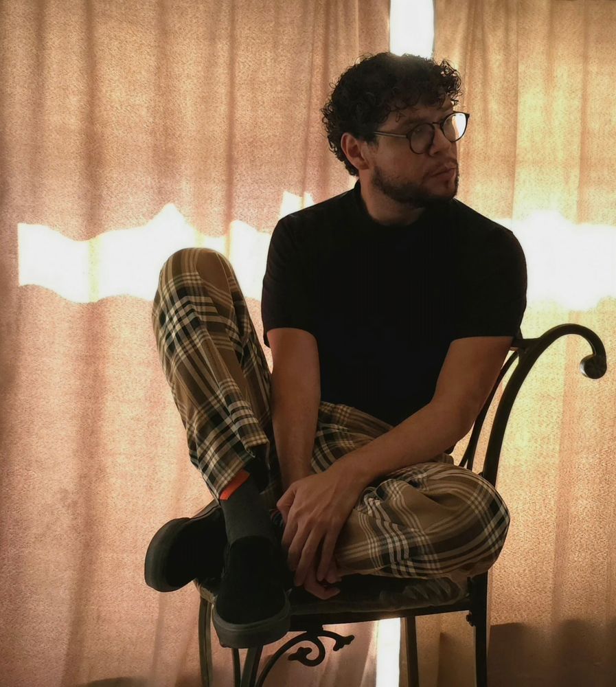 I'm sitting on a chair wearing striped pants, black shoes, black shirt, and glasses. I'm looking off to the side while curtains hang behind me with a streak of light running through them. The lighting is all very brown.