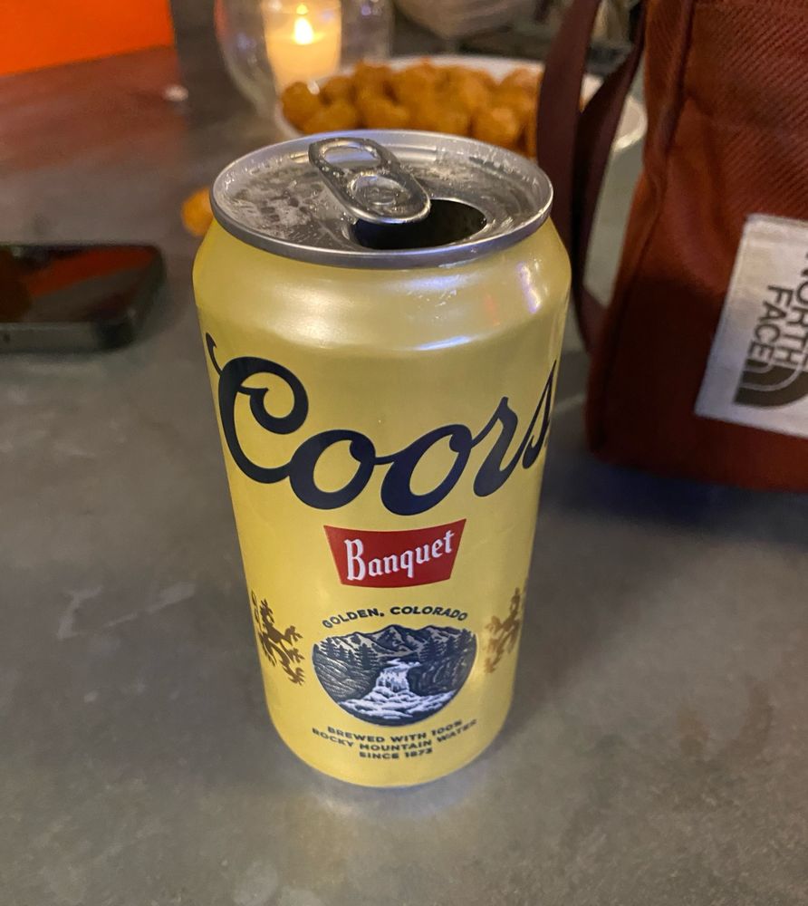 A can of Coors Banquet on a steel table with a plate of tater tots in the background