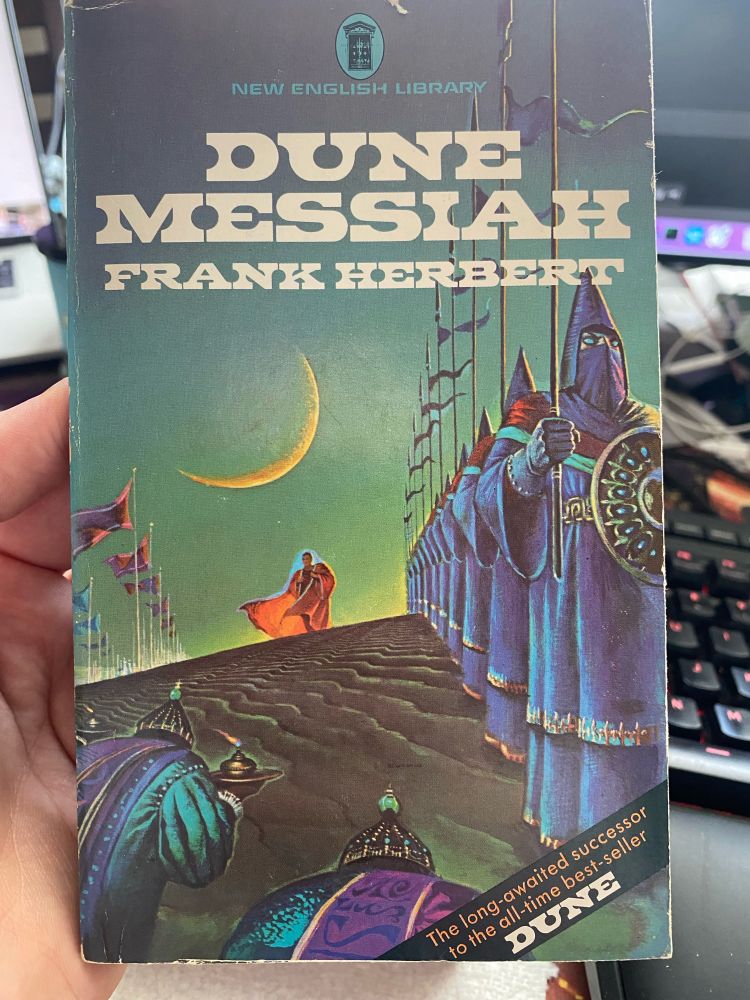 The cover of a UK edition of Dune Messiah from the 1970s. A cloaked figure is approaching between lines of hooded and masked figures holding polearms and shields under a crescent moon.
