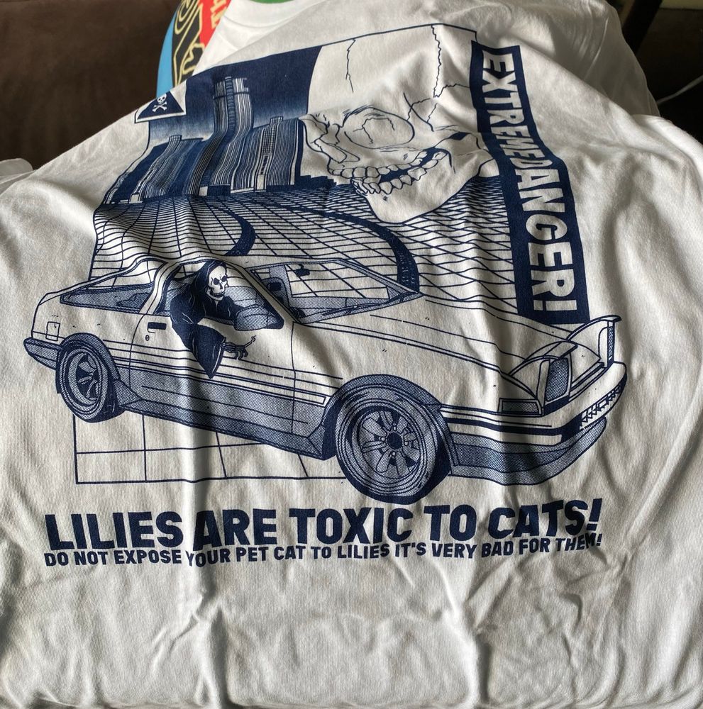 A shirt with art of the grim reaper doing a power slide in a Toyota AE86 in front of the Renaissance Center and a large skull in the corner with the text: Exteme Danger! Lilies are toxic to cats! Do not expose your pet cat to lilies it’s very bad for them! 