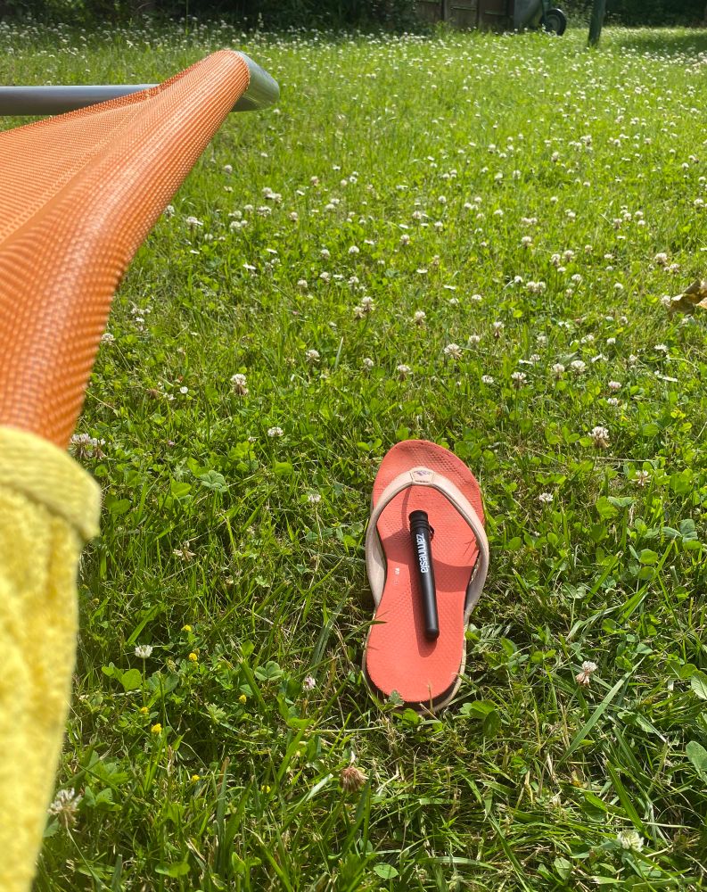 auf einer Wiese neben orangefarbener Saunaliege mit gelbem Handtuch steht orangefarbener Flipflop auf dem eine schwarze Hülse von Zamnesia liegt