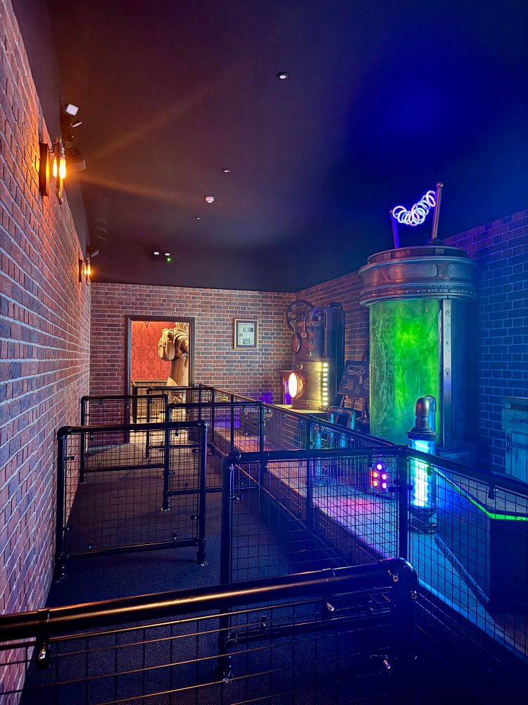 An indoor queue room with empty queue barriers on the left and theming elements such as vats and coils on the right. 