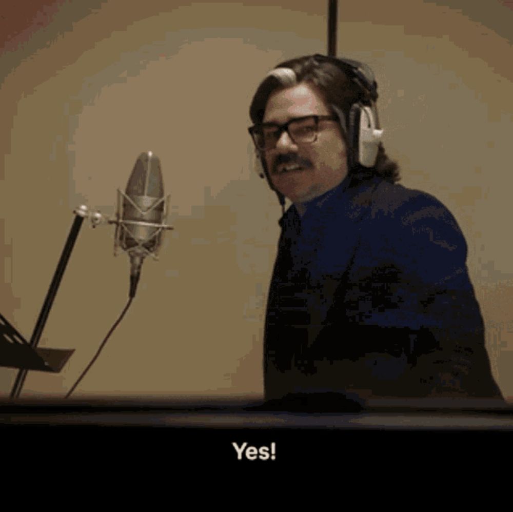 A man in glasses and headphones stands in front of a microphone with the word "yes!" subtitled below him