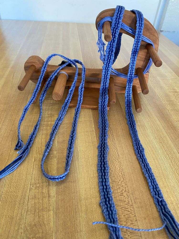 Woven straps draped over a small inkle loom on a butcher block surface.