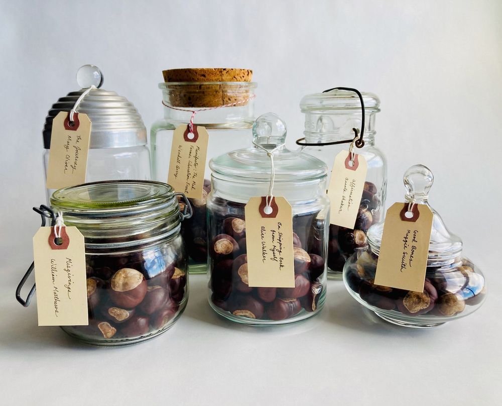 six glass jars of different shapes and sizes, each containing buckeyes with words written on them. Each jar has a tag with the title and author of a poem.