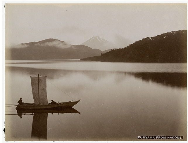A photo in grayscale, of a small boat on the water with mountains in the background.