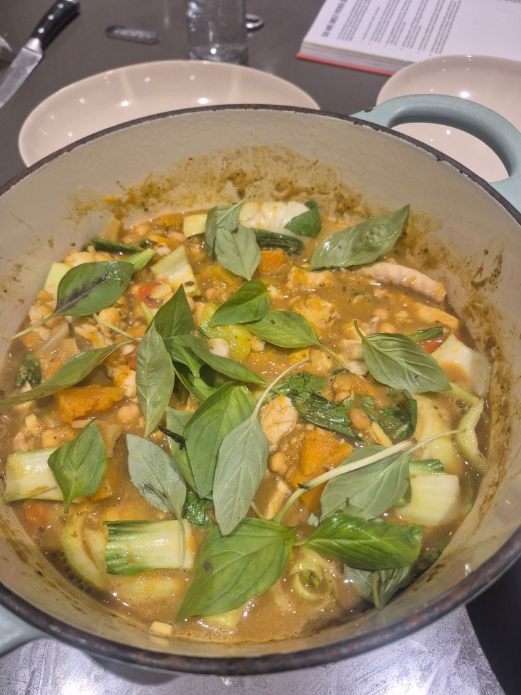 A casserole dish full of curry made with sweet potato, onions, garlic, ginger, green Thai curry paste, coconut milk, pak choice, cod, chickpeas and garnished with Thai Basil.