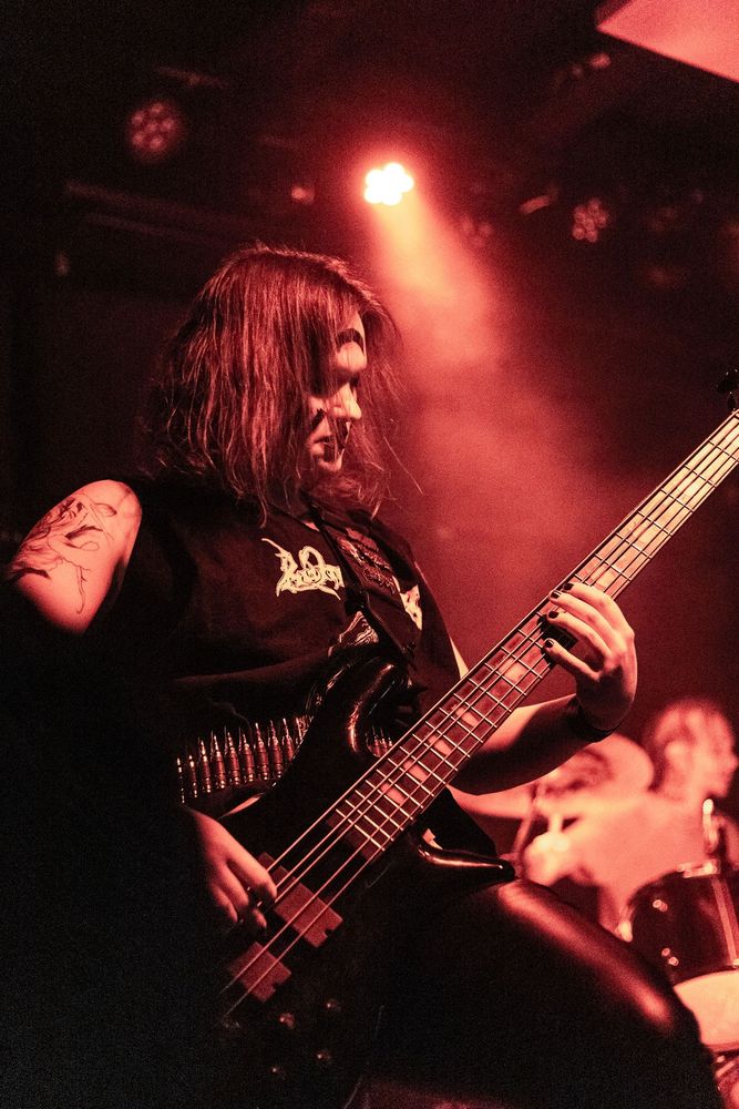 A woman playing bass on stage she's bathed in red light playing a black Spector bass wearing leather pants and a  Runemagick shirt belted at her waist with a bullet belt 
