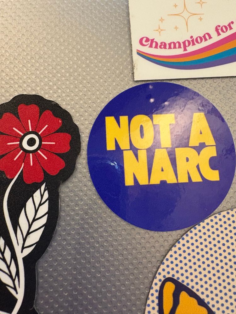 Laptop case with Not a Narc sticker, “Harm reduction build community” sticker, and partial views of 2 other stickers 