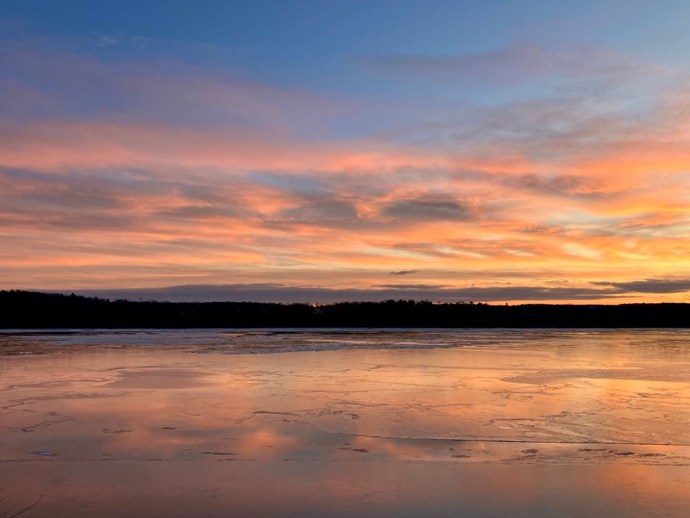 A serene landscape featuring a frozen body of water reflecting colorful clouds at sunset, with soft hues of orange, pink, and blue in the sky. The silhouette of trees is visible in the background.