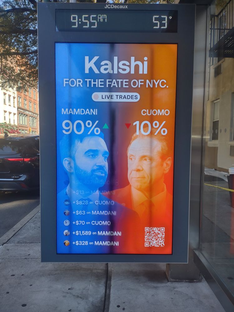 Photo of a dynamic bill board at a bus stop on a city street sidewalk with a black steel frame with a black time and temperature screen across the top and a colored rectangular screen filling most of the photo. The colored screen has a blue and orange background and reads Kalshi in big letters at the top, "for the fate of NYC" under that and a lozenge that says live trades. Beneath that, the screen is split with Mamdani on the left at 90% and Cuomo on the right with 10% and a feed of people placing dollar bets scrolling vertically at the lower left. There is a white Q R code in the lower right 