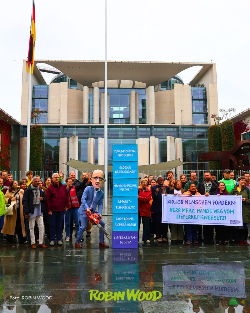 Aktionfoto mit vielen Menschen vor dem Bundeskanzleramt in Berlin, ein als Merz verkleideter Aktivist mit Kettensäge, Tafeln mit diversen Forderungen sowie Transpi: “210438 Menschen fordern: Herr Merz, Hände weg vom Lieferkettengesetz!”