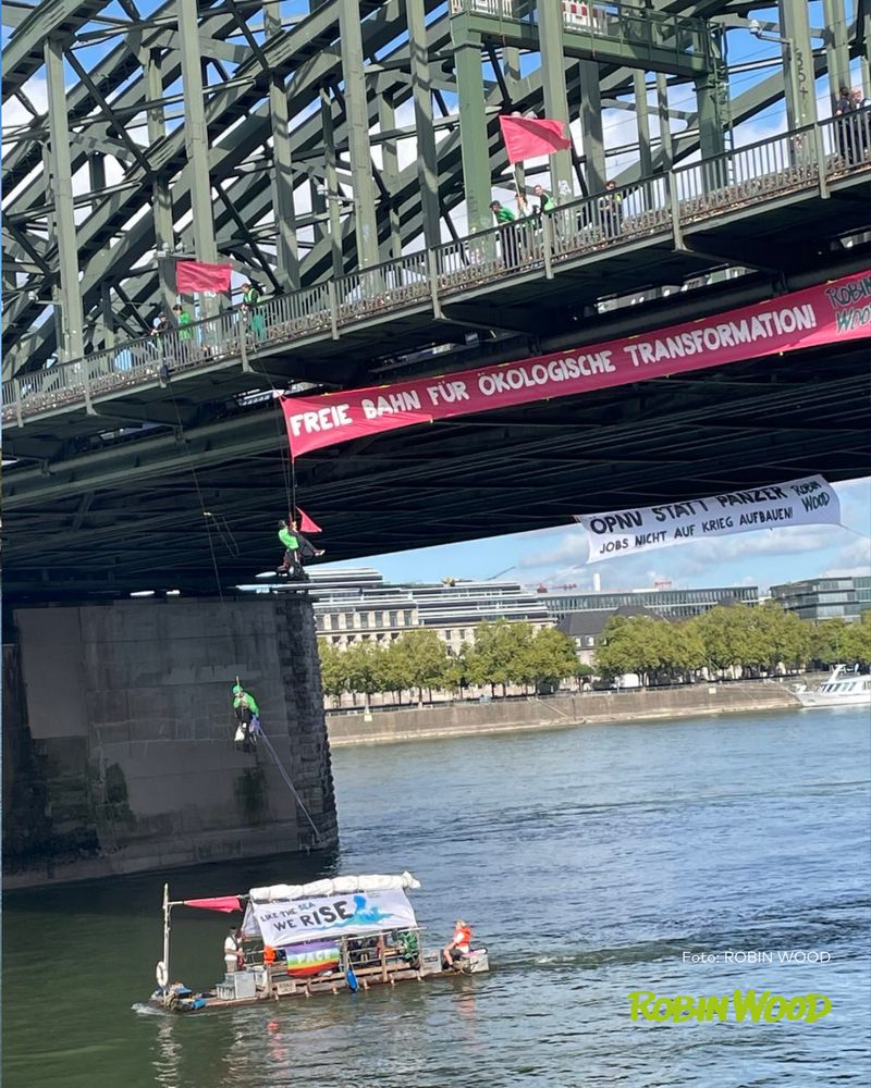 Aktionsfoto Köln Hohenzollernbrücke, großes Transparent mit der Aufschrift „Freie Bahn für ökologische Transformation!“ und darunter kleineres Transpi „ÖPNV statt Panzer – Jobs nicht auf Krieg aufbauen.“ dabei mehrere Kletter*innen, darunter auf dem Rhein das ROBIN WOOD-Floß

