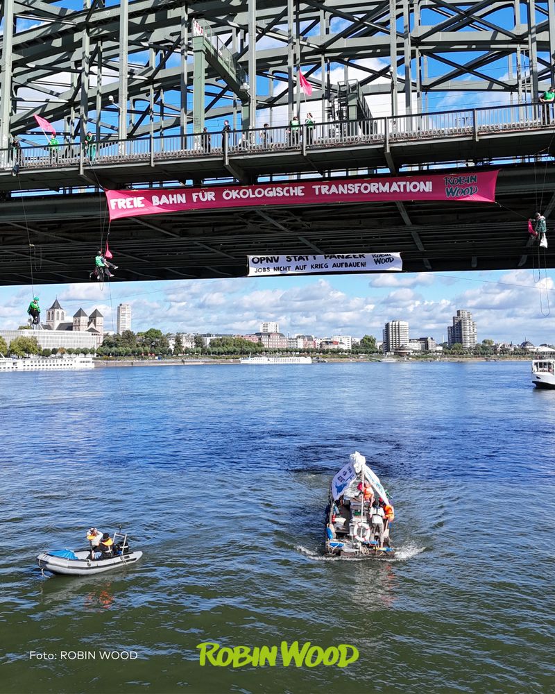 Aktionsfoto Köln Hohenzollernbrücke, großes Transparent mit der Aufschrift „Freie Bahn für ökologische Transformation!“ und darunter kleineres Transpi „ÖPNV statt Panzer – Jobs nicht auf Krieg aufbauen.“ dabei mehrere Kletter*innen, darunter auf dem Rhein das ROBIN WOOD-Floß
