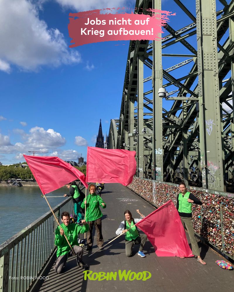 Mehrere Aktive in ROBIN WOOD-Aktionskleidung auf der Hohenzollernbrücke in Köln mit pinkroten Flaggen in den Händen, Schrift: “Jobs nicht auf Krieg aufbauen”