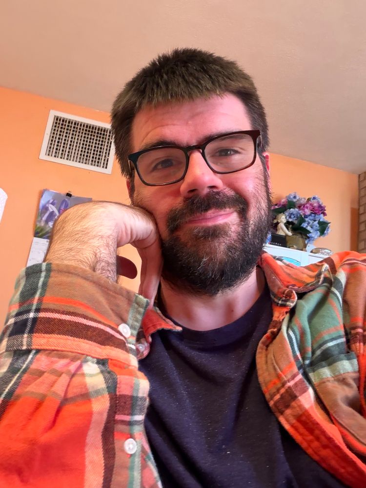 Bridge leaning on his arm in a selfie in a purple t shirt and orange and green flannel shirt. 
