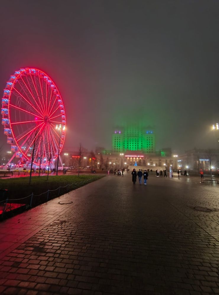 On a foggy night, a big wheel grows red and the Palace of Culture and Science grows green, creating a somewhat spooky atmosphere.