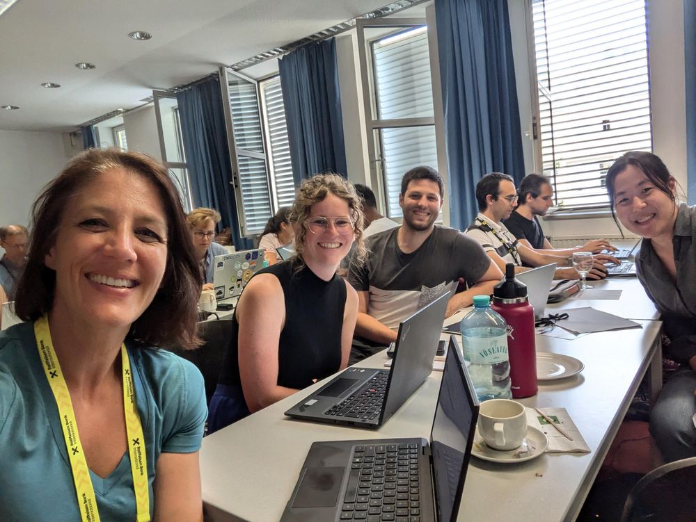 Participants at an R Dev Day sat at desks in classroom layout. The closest group has turned to smile at the camera, while others further away are glued to their laptop. A mix of ages, genders and nationalities are represented.