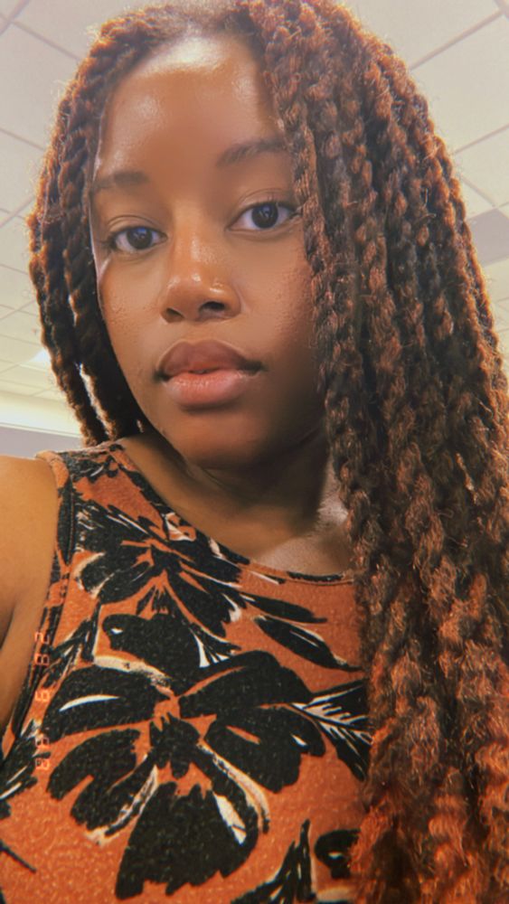 Bluesky User MadamZeti taking a selfie. She is wearing a burnt orange and black sleeveless dress and her hair is in brown/black Marley twists