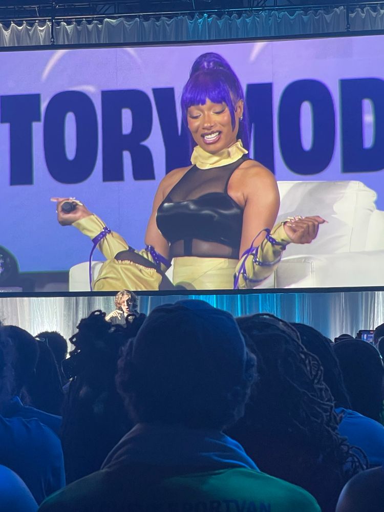 Megan Thee Stallion at DreamCon cosplaying as Yoruichi from Bleach. She is wearing a black mesh bodysuit, beige accessories around her neck, arms, and legs and has a purple high pony tail
